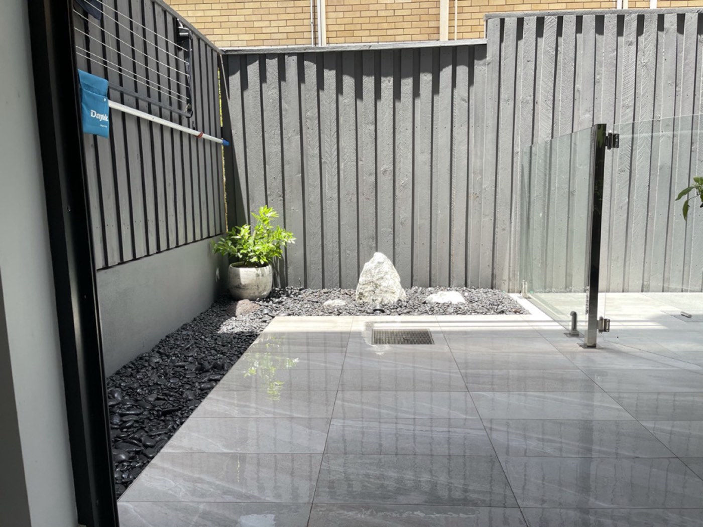 A small modern courtyard with grey porcelain paving, a feature rock and a black pebble border against a dark fence